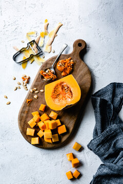 A sliced butternut squash with seeds and cubes on a wooden cutting board, alongside a peeler and a dark linen cloth.