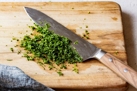 A kitchen knife with a wooden handle is placed on a wooden cutting board next to freshly chopped green herbs.