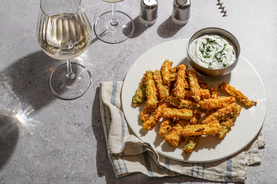 Healthy crunchy zucchini nuggets with sauce, on a grey stone background with bright sunlight, and a glass of white wine