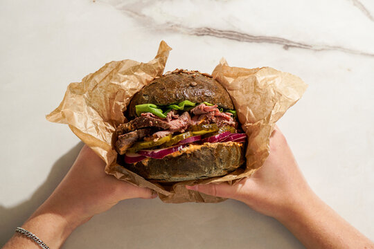 gourmet Roast Beef Sandwich, fresh lettuce, pickles, red onion, held person's hands, with a neutral background and hash sun lighting