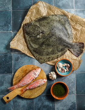 turbot flounder on a rustic wooden cutting board with olive oil, garlic, and green sauce on a tile background