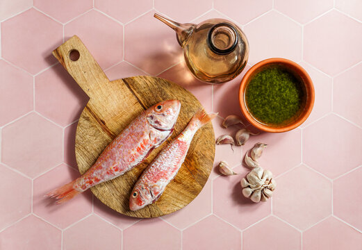 resh red mullets on a rustic wooden cutting board with olive oil, garlic, and green sauce on a pink hexagonal tile background