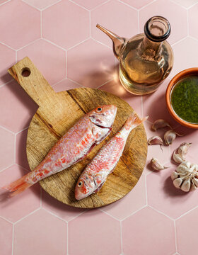 resh red mullets on a rustic wooden cutting board with olive oil, garlic, and green sauce on a pink hexagonal tile background