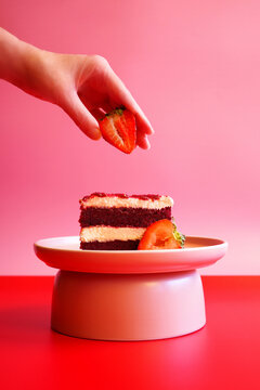 Female hand placing strawberry on top of slice of double layer red velvet cake. Pop art food color blocking aesthetic.