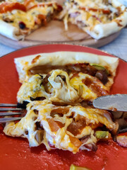 Appetizing slice of delicious pizza in red plate against background of sliced ​​pizza served on wooden round tray. Delicious and healthy food. Close-up. Selective focus.