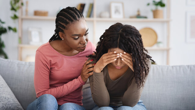 Attentive african american lady comforting her upset crying girlfriend or sister, giving her hug, saying supportive words, living room interior, copy space. Support in friendship concept