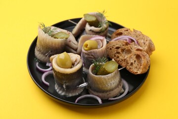 Delicious herring rolls with olives, pickles, dill, onion and bread pieces on yellow table, closeup