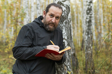 A man with a beard is writing in a red notebook while standing next to a tree