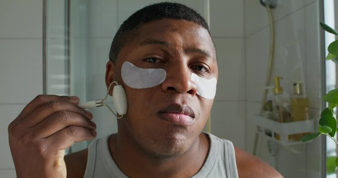 A man with eye patches is doing a facial massage with roller massager. He looks at the camera like it's a mirror. Close-up of his face