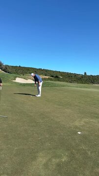 Man putting on a green golf course beside the sea under a clear blue sky. The ball narrowly misses the hole. Coastal landscape in the background.
