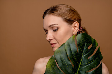 Beautiful young woman middle age lady with clean fresh skin touching face in tropical green leaf closed eyes posing standing isolated in beige background. Cosmetology beauty and spa care concept.