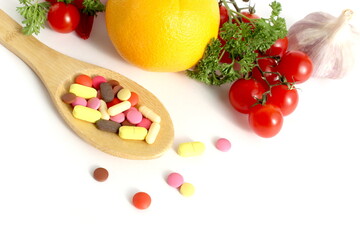  On a white background lies a wooden spoon with medicine and in the background are vegetables and fruits.