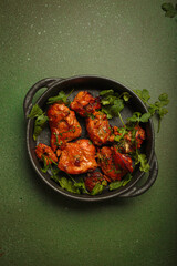 Top view of tandoori chicken, an Indian dish of roasted marinated chicken chunks in spiced curry sauce, served in a cast iron pan on a green background and garnished with fresh coriander leaves