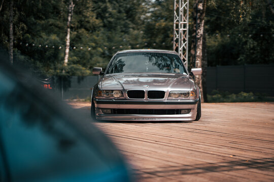 Static shot of BMW E38 7 Series on air suspension with polished wheels at outdoor car show &ndash; slammed luxury sedan displayed on wooden platform under warm daylight with show cars in the background