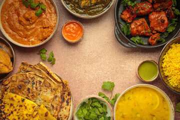 Overhead view of a variety of Indian dishes on a beige table, including curry, tandoori chicken, chicken tikka masala, dal, Palak paneer, paratha, rice, samosas. Cuisine of India, copy space