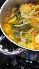 Traditional Portuguese codfish stew, known as caldeirada de bacalhau, prepared in a stainless steel pot with sliced potatoes, onions, tomatoes, peppers, herbs and olive oil.Homemade meal on a stovetop