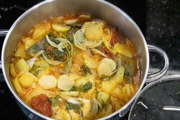 Traditional Portuguese codfish stew, known as caldeirada de bacalhau, prepared in a stainless steel pot with sliced potatoes, onions, tomatoes, peppers, herbs and olive oil.Homemade meal on a stovetop