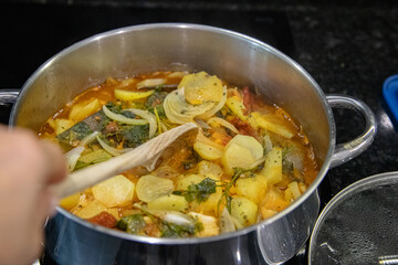 Traditional Portuguese codfish stew, known as caldeirada de bacalhau, prepared in a stainless steel pot with sliced potatoes, onions, tomatoes, peppers, herbs and olive oil.Homemade meal on a stovetop