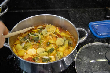 Traditional Portuguese codfish stew, known as caldeirada de bacalhau, prepared in a stainless steel pot with sliced potatoes, onions, tomatoes, peppers, herbs and olive oil.Homemade meal on a stovetop