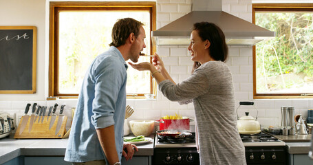 Couple, cooking and feeding food in kitchen at house for organic ingredients, meal prep and healthy lunch. Woman, man and taste recipe for nutrition salad, vegan diet and romance for bonding together