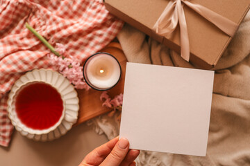 Hand holding a blank square card with a cozy spring setting of tea, a lit candle, pink flowers, and a gift box in the background, perfect for a greeting or invitation mockup