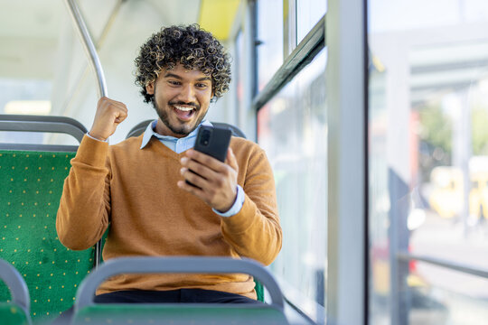 A young man on a bus is happily looking at his phone, celebrating good news. He is holding his fist up in excitement with a big smile.