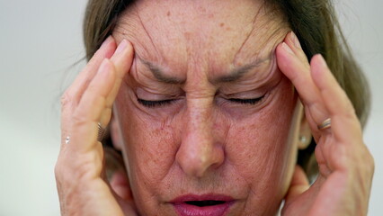 Close-up of senior woman with eyes tightly shut, pressing fingers to temples, intense focus on pain...