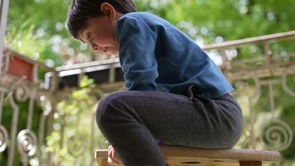 Close-up of a boy sitting on a wooden stool outdoors, hands gripping the edges, surrounded by lush...