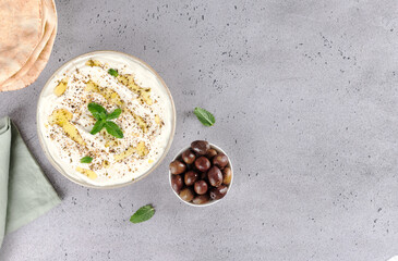 Bowl of labneh cheese with olive oil, zaatar, fresh mint, olives, pita bread on grey background, Top view. Copy space.