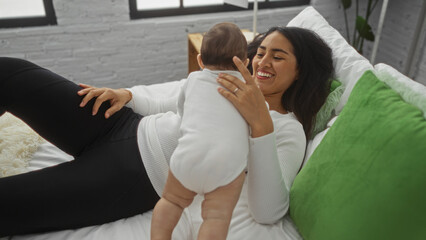 Mother enjoying quality time with baby girl on bed in cozy home bedroom, displaying love and warmth in an indoor family setting with soft green accents.