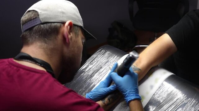 Top-down view of a tattoo artist using a tattoo machine during a session. The artist is focused on the process, wearing gloves. Tattooing in progress. 