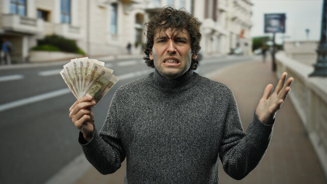 Hispanic man holding russian rubles outdoors on a city street, expressing confusion or frustration in a public urban setting with a backdrop of buildings.