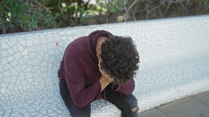 Young man sitting in outdoor urban park with head in hands showing stress or sadness, surrounded by city environment, casual outfit suggesting a moment of reflection.