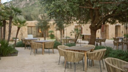Blurred image of an outdoor mediterranean restaurant terrace with wicker chairs and tables under trees, creating a peaceful and cozy ambiance.