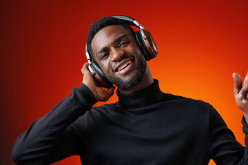 Young man enjoying music with headphones, wearing a black turtleneck against an orange background,...