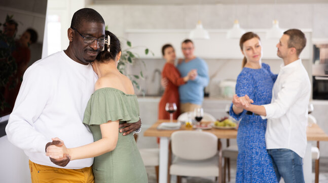 Smiling black man in casual white sweater tenderly embracing woman in green off-shoulder dress, enjoying slow dance at cozy friendly house party - Powered by Adobe