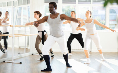 Obraz premium Focused middle aged African American man confidently leading ballet class with male and female amateur dancers following his movements in spacious sunlit dance studio