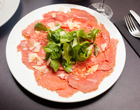 Thin slices of meat carpaccio, cheese and arugula on white plate served on table in restaurant. Traditional Italian dish