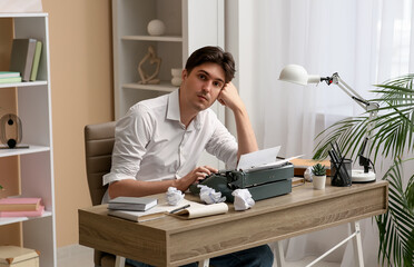 Young writer with vintage typewriter and crumpled paper sitting at table in room