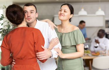 Fototapeta premium Positive young man with wife warmly hosting and kissing on cheek female guest arriving for friendly dinner gathering at home