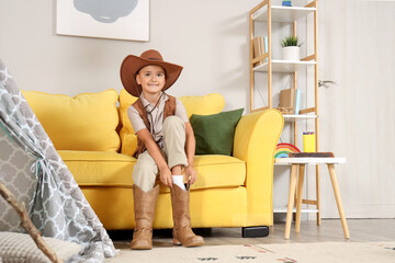 Fototapeta premium Smiling young boy in cowboy hat and boots sitting on yellow sofa, child's playroom adventure