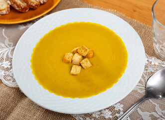 There is dinner plate with delicate cream soup of yellow color on table. Carrot mousse is decorated with small toasted crispy croutons