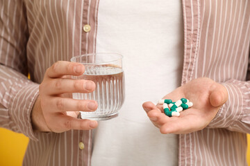 Young man with heap of pills and glass of water on yellow background, closeup