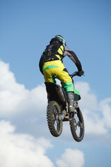 Rear view of a motocross rider in bright gear performing a jump on an off-road motorcycle against a blue sky with clouds