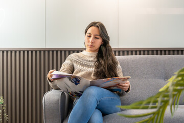 A young woman is sitting comfortably on a modern grey sofa in what appears to be a clinic or office waiting room with a magazine