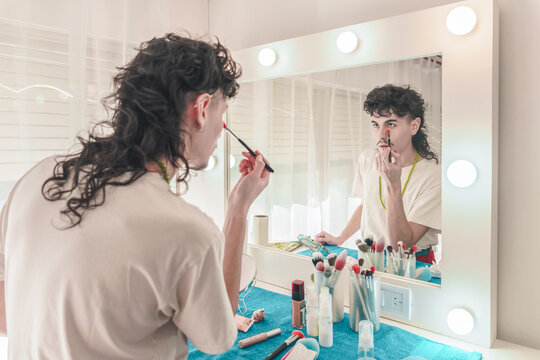 Young non-binary person applying makeup in front of a mirror with light bulbs