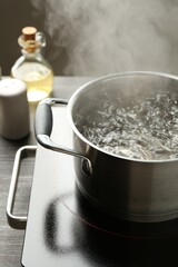 Cooking pot with boiling water and stove on wooden table against grey background, closeup