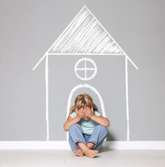 Orphanage. Sad little girl on floor near grey wall with drawing of house