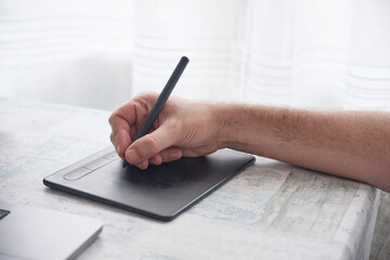 Close-up of an unrecognizable man’s hand using a wireless drawing tablet with a stylus pen beside...