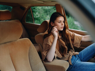 Thoughtful young woman driving in a vintage car, capturing emotions of nostalgia and adventure in nature. Stylish brown outfit and serene greenery create a tranquil atmosphere.